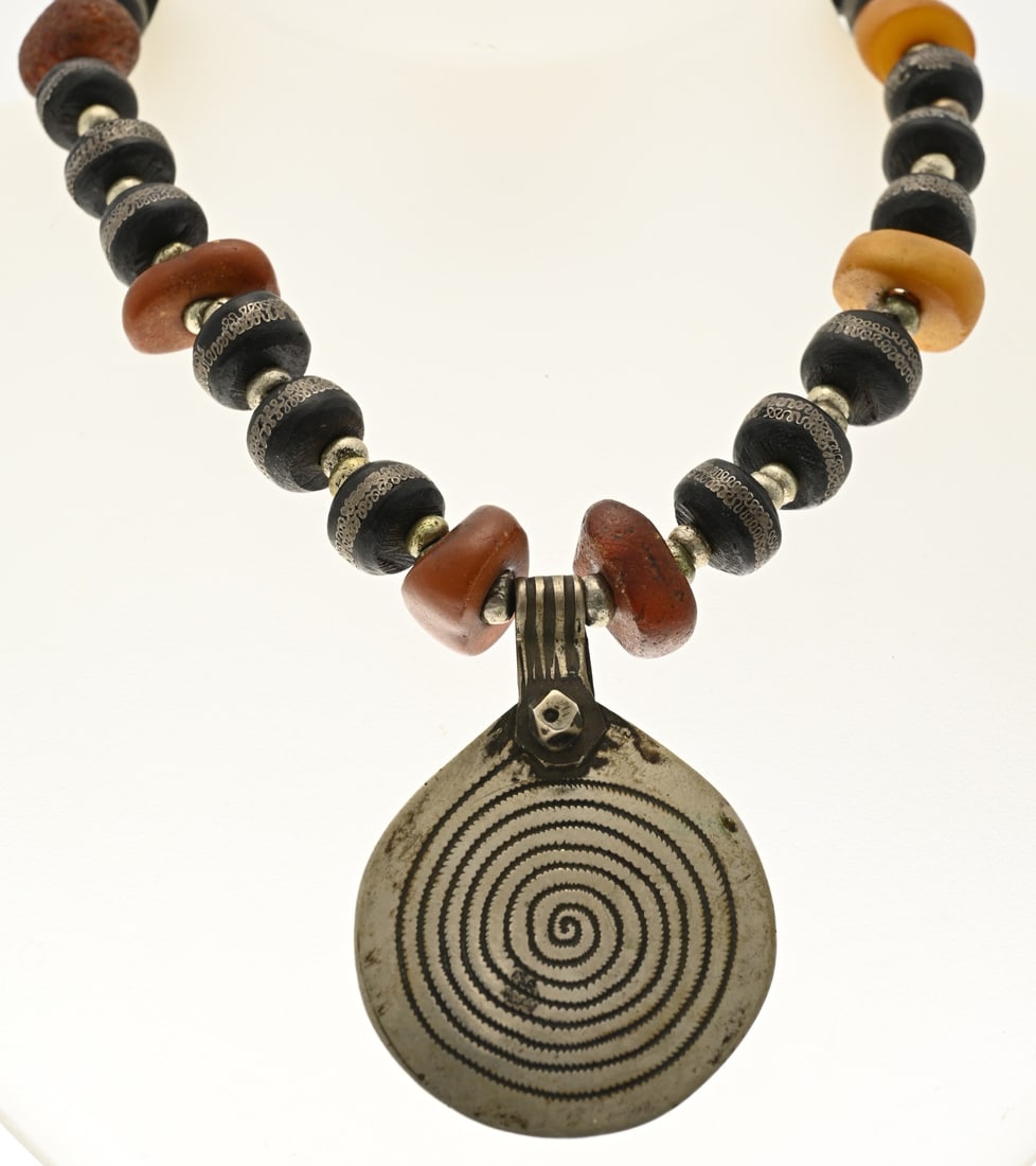 Berber necklace: Berber necklace with black and amber beads decorated with a white metal element with spiral detailing. With hook clasp. Length 49 cm. Pendant size ø 53 mm. In good condition.