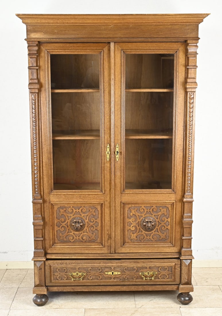 German bookcase, 1880: German Gründerzeit oak book/display cabinet with carved lion heads and a drawer. Circa 1880. Dimensions: 181 x 55 x 112 cm. In good condition.