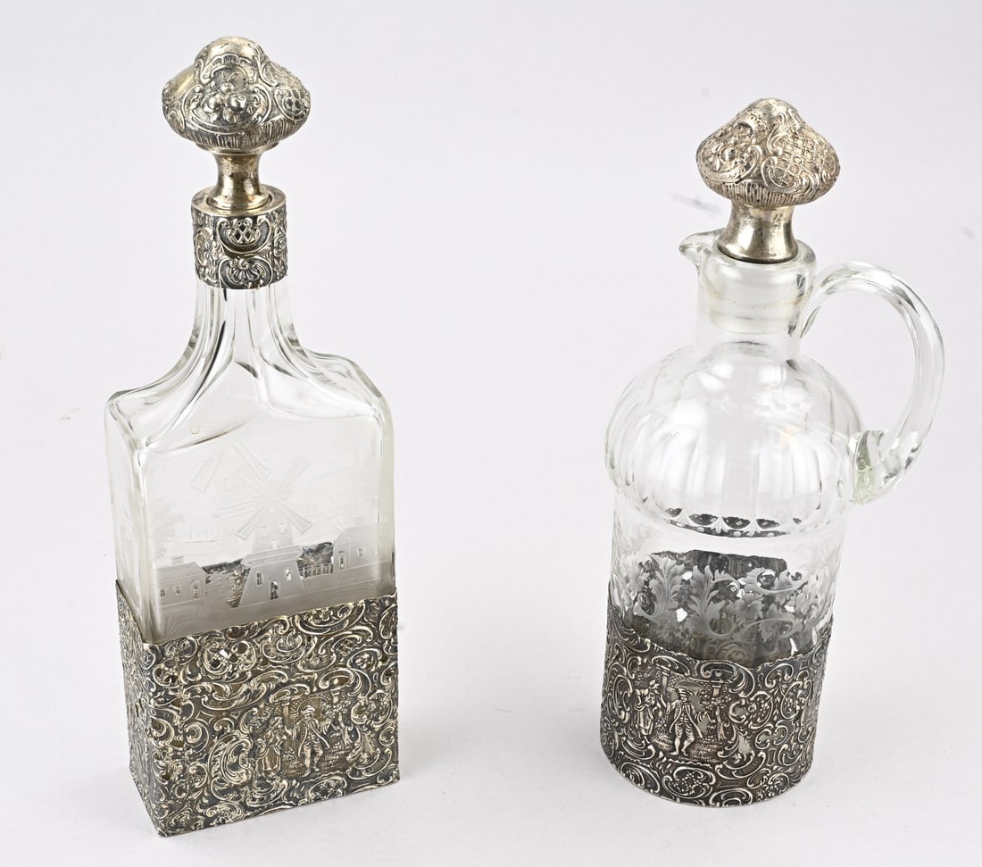 2 Decanters with silverware: Two crystal decanters with silver, 800/000. A rectangular decanter decorated with etched motifs of windmills, houses, and ships. The base and collar feature openwork silver with floral decoration and