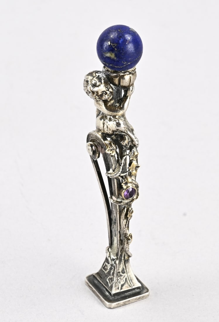 Silver stamp: Silver stamp with putti holding a lapis lazuli globe. Decorated with gilded leaf elements and a smooth amethyst setting. 8x1.3x1.5cm. Lapis sphere broken, glued. Approx. 25 grams. In good condition.