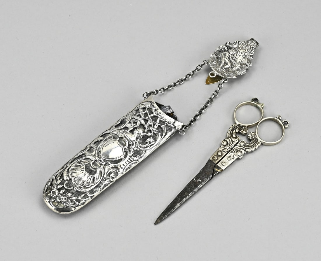 Scissors in travel case: Antique case with silver detailing and a skirt hook decorated with figures and floral ornamentation. Inside the case is a pair of scissors with silver handles featuring volutes and floral