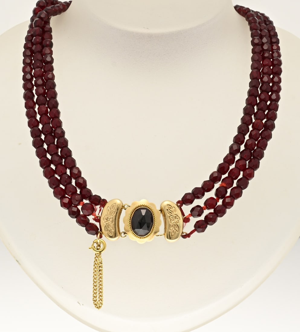 Garnet necklace with gold clasp: Necklace of 3 rows of glass garnets, ø 5 mm., attached to an oval yellow gold clasp, 585/000, with a scalloped edge and set with a garnet stone. The clasp features 2 engraved gold end pieces and