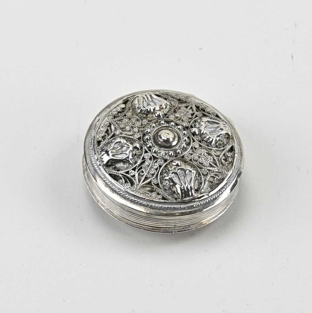 Round silver box: Silver pillbox, 833/000, round model with ribbed decoration. The lid features filigree decoration with 4 crown-shaped and floral elements. Approx. 32 grams. ø 62 mm. MT.: unclear, Schoonhoven.