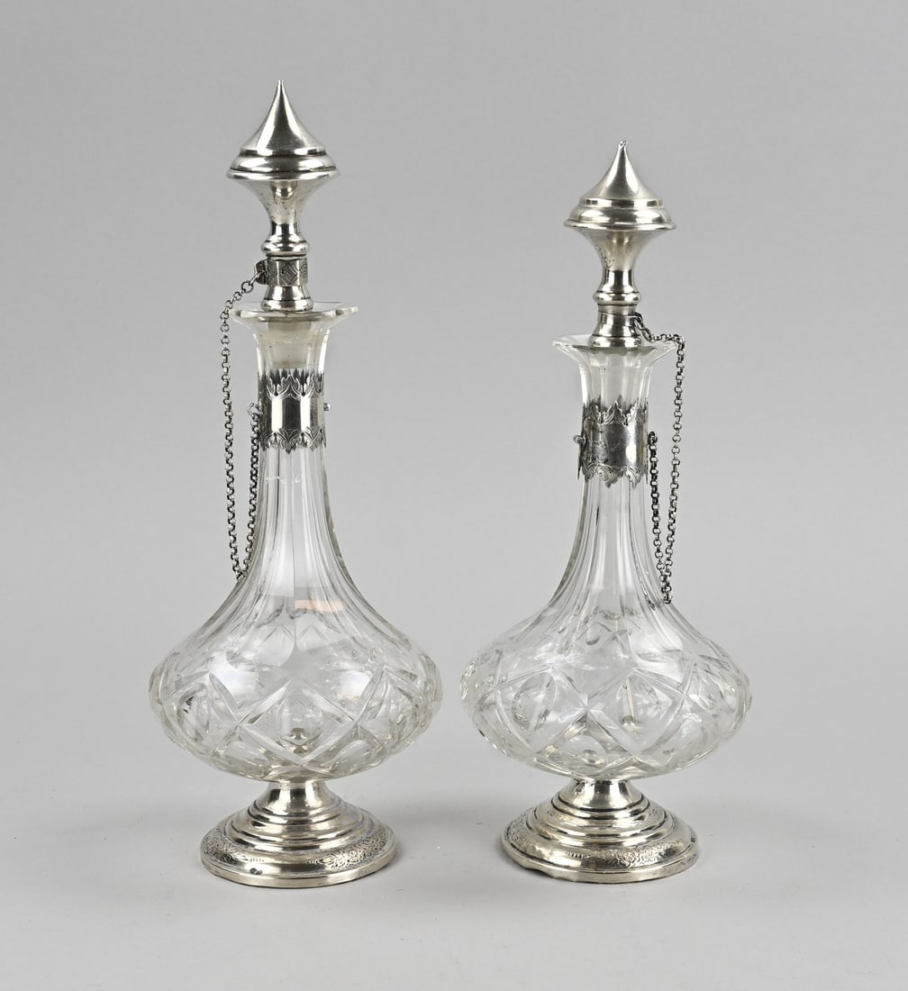 2 Decanters with silverware: Two small crystal decanters with silver, 833/000. Decanters with cut work, placed on a round silver base with engraving. A silver collar with engraving, a chain, and a silver stopper. ø10.5x28cm. Sto