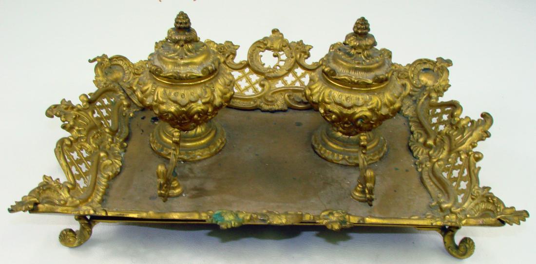 ELABORATELY CAST DOUBLE INKWELL: Possibly gilded cast bronze double inkwell with pen holder; hinged inkwells retain porcelain inserts; footed openwork gallery base; floral and scrollwork motif; some areas of greenish patination / dis
