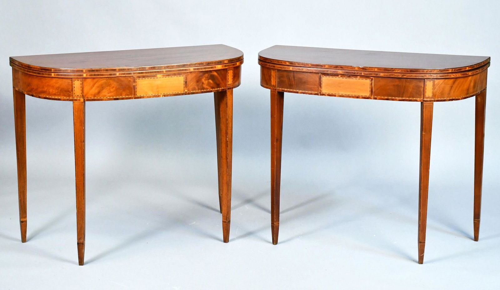 A PAIR OF INLAID MAHOGANY CARD TABLES.: A pair of demi lune tables each with a mahogany top and conforming apron with satinwood inlay panels, checkered and other inlays, on line inlaid double tapered legs. One table was made later to match