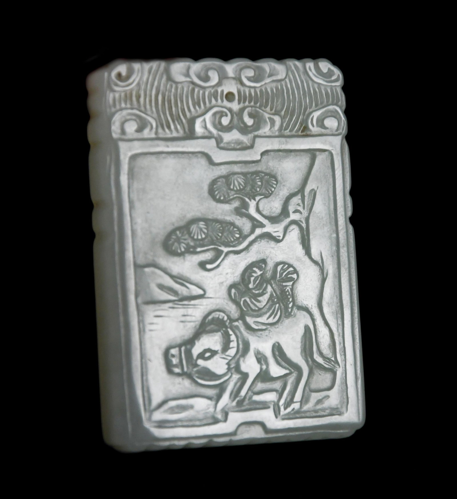 CARVED CELADON JADE PENDANT.: A carved pale celadon jade rectangular pendant, one side with characters, other side with image of figure on the back of an ox. 2.5” x 1.75”.