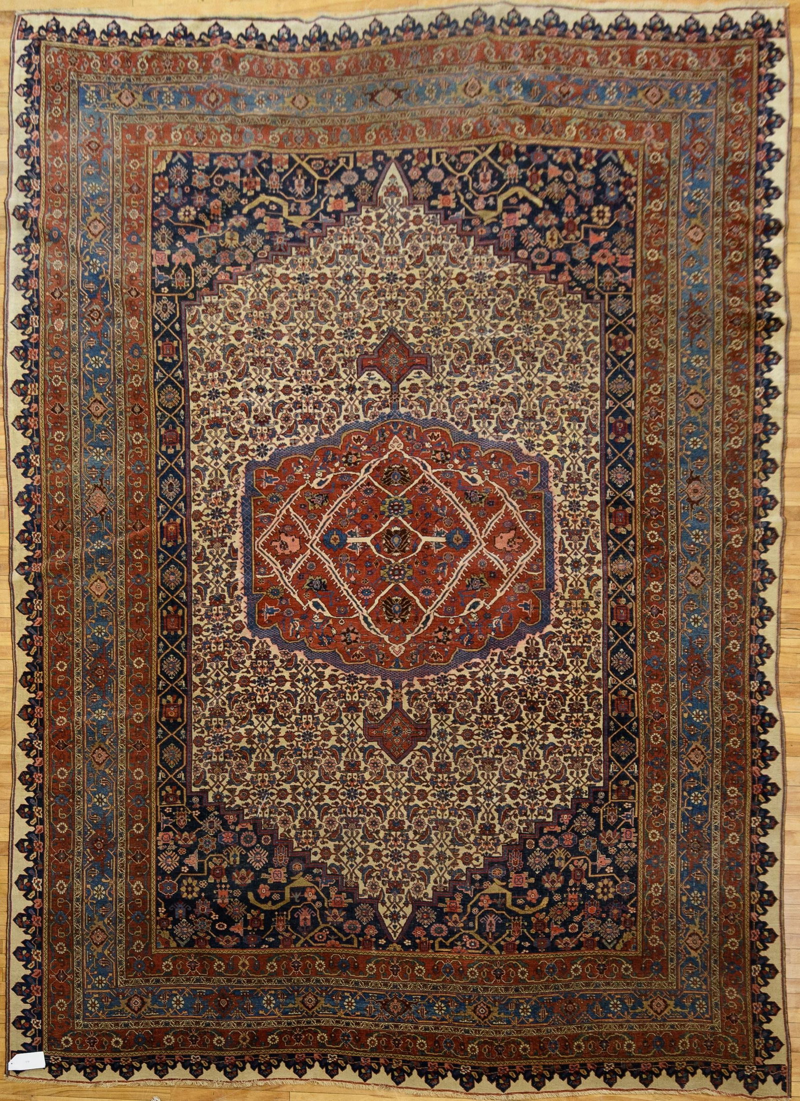 ANTIQUE PERSIAN IVORY GROUND ORIENTAL RUG.: A ca. 1920 antique Persian oriental rug, the ivory field with an allover herati design centered by a stepped rust and blue medallion with pendants, flanked by indigo corner spandrels, within a rust pa