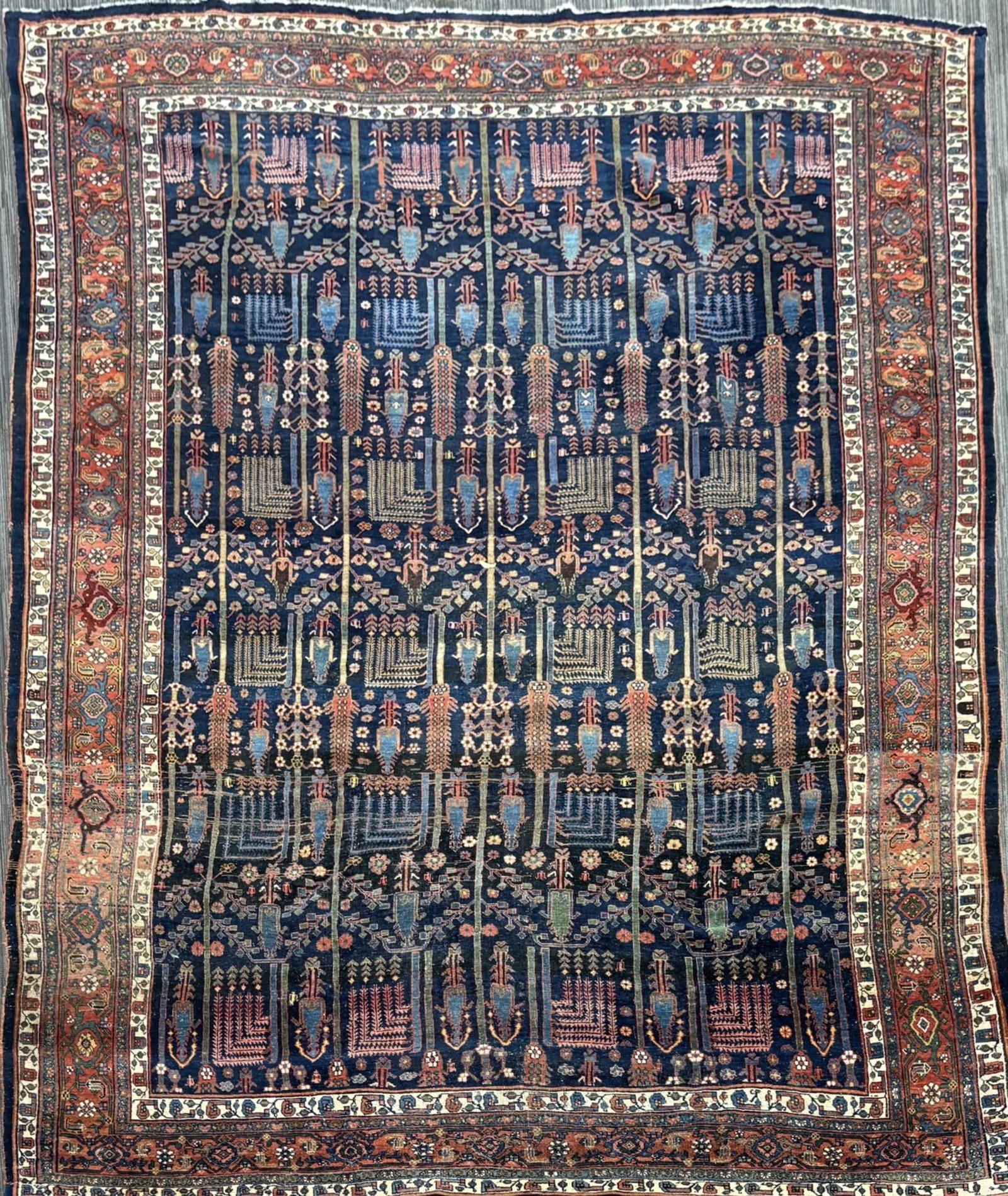 ANTIQUE PERSIAN RUG, TREE OF LIFE PATTERN.: An antique Persian room size rug, featuring a highly decorative garden or “tree-of-life” design arranged in vertical rows across a deep navy field. The field is populated with stylized cyp