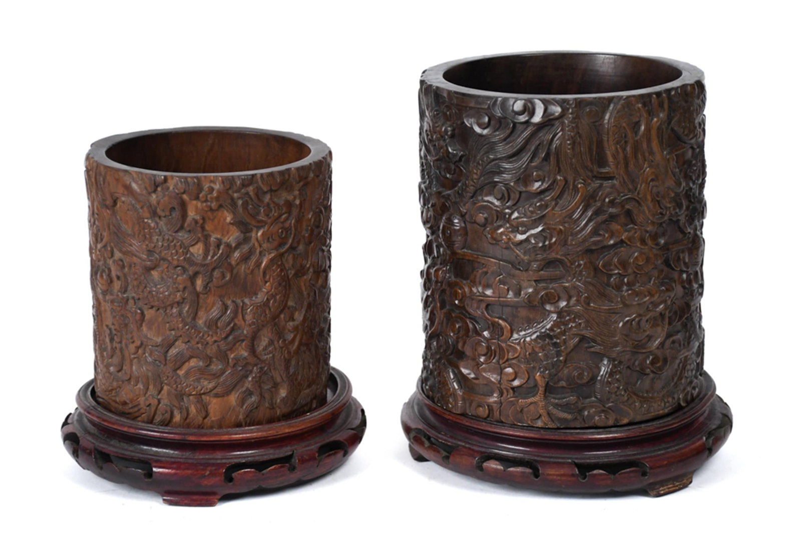 TWO ANTIQUE CHINESE CARVED HARDWOOD DRAGON BRUSH POTS: Two antique Chinese carved hardwood brush pots (bitong), each featuring deeply carved all-over relief decoration of writhing dragons amid clouds and crashing waves, each on a carved wood stand,