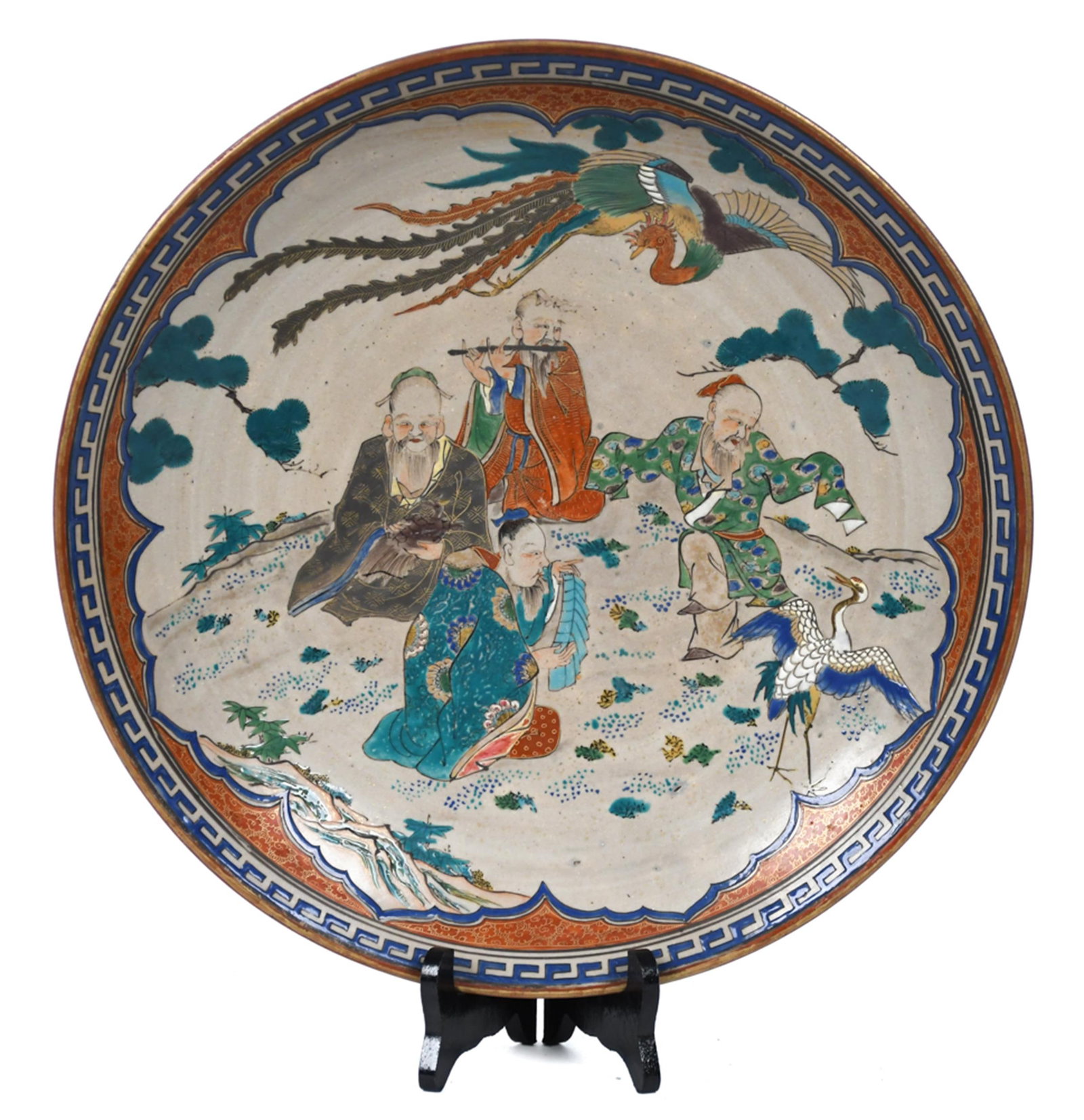 ANTIQUE KUTANI PORCELAIN CHARGER.: An antique Japanese Kutani porcelain charger, the interior painted in polychrome enamels with a scene of sages, a crane, and a phoenix amid pine trees, within a shaped white reserve. A rust-red and co