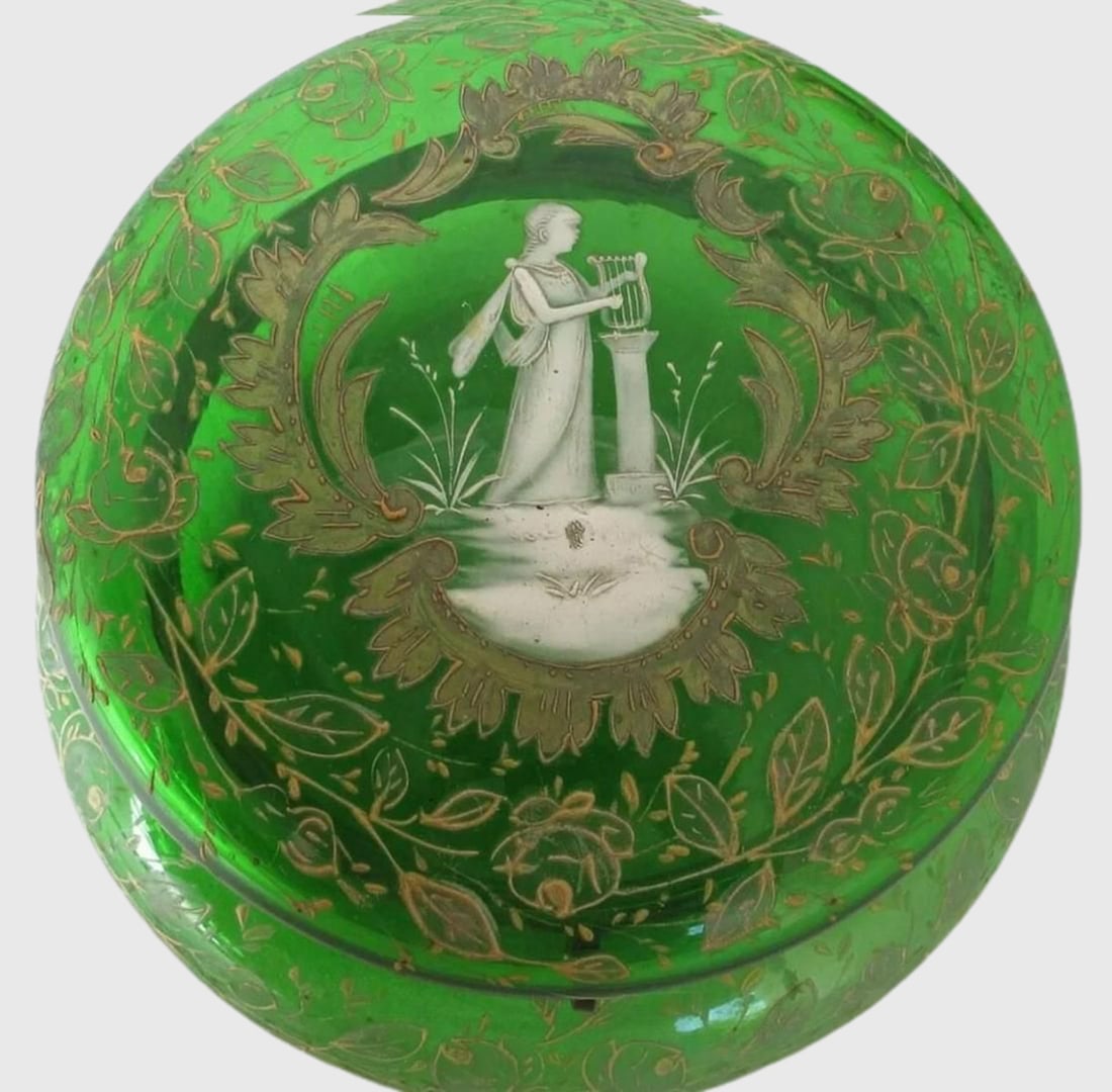 BOHEMIAN MARY GREGORY GREEN GLASS DRESSER TRINKET WITH: Description: Bohemian Mary Gregory Green Glass Dresser Trinket with white enameled. Mary Gregory lid picturing a classic woman playing a lyre. Size: 6" Diameter and 3 1/2" Tall