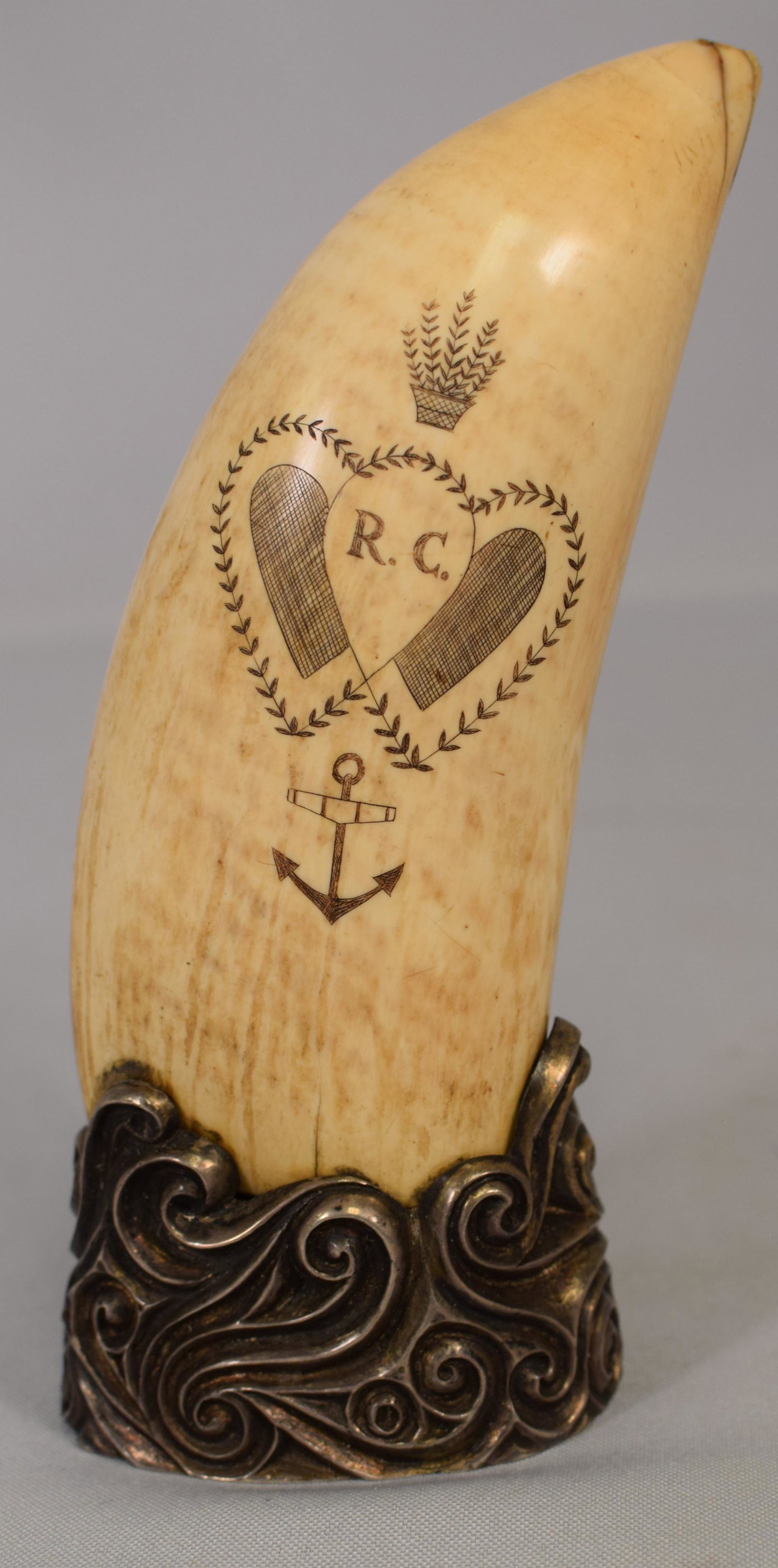 EARLY STERLING MOUNT SCRIMSHAW WHALE TOOTH: Large early whale tooth with scrimshaw romantic heart motif design. Sterling silver scroll "waves" mount. Marked R.C. with arrow. Measures approximately 6. Approximately 638 grams,overall. Some loss t