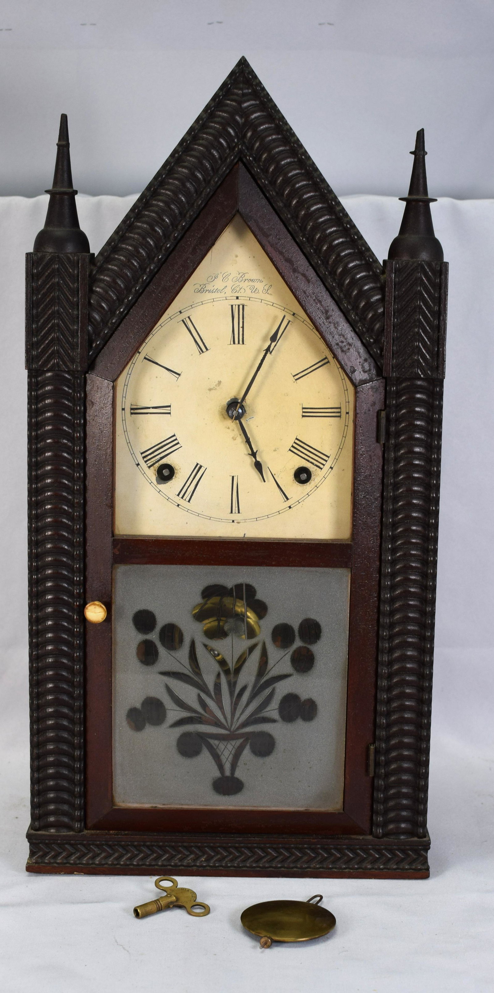 J.C. BROWN RIPPLE FRONT STEEPLE CLOCK: Ca 1850. Deeply carved mahogany ripple front steeple clock. Painted metal face with Roman Numerals (some loss and discoloration). Signed J.C. Brown, Bristol, Ct. USA. Eight day time andstrike, wire go