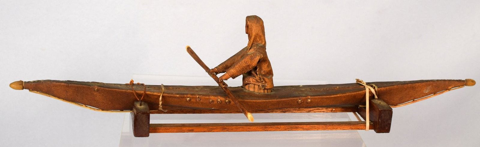 EARLY ESKIMO SEALSKIN MODEL KAYAK WITH STAND: Fine old patina Eskimo seal skin model kayak. 16 1/2 inches long. Hand made. Figure made of seal skin with carved wooden face. Paddle with bone tips. End of kayak with carved horn tips. Woodenbase. So