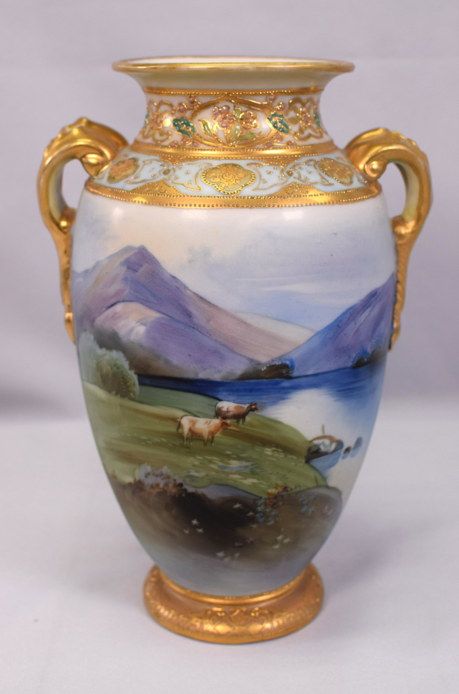 JAPANESE HANDPAINTED NIPPON PORCELAIN VASE: Scenic landscape with cows and gilt handles and accents. Grazing cows with lake at foothills of mountains. Nippon Morimura (Japanese 1911-1921). 10 inches tall x 6 1/2 handle to handle x 5inches. Ship
