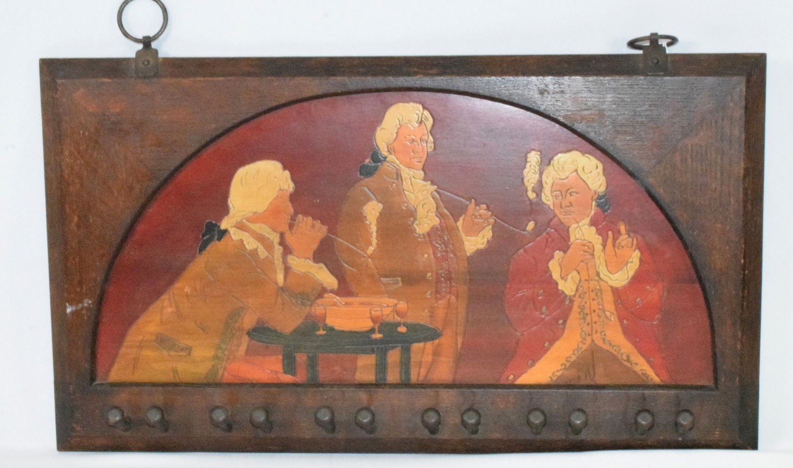 FLEMISH ART HANGING PLAQUE: Incised plaque with detailed painted Colonial tavern scene. Three gentlemen with white powder wigs, clay pipes and wine goblets. Oak frame with 12 pegs for hanging hats or coats, iron rings.Measures 1