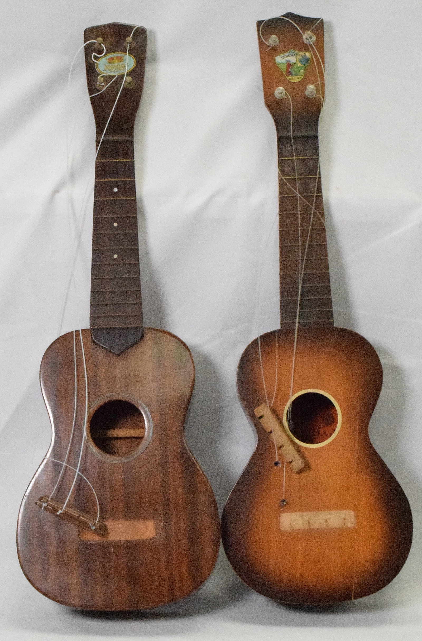 2 VINTAGE UKULELES: 1) Marked with label The Serenader. Approx. 21 inches long. Crack on front. Loose strings. 2) Label Regal. 21 inches long. Mahogany. Loose strings.