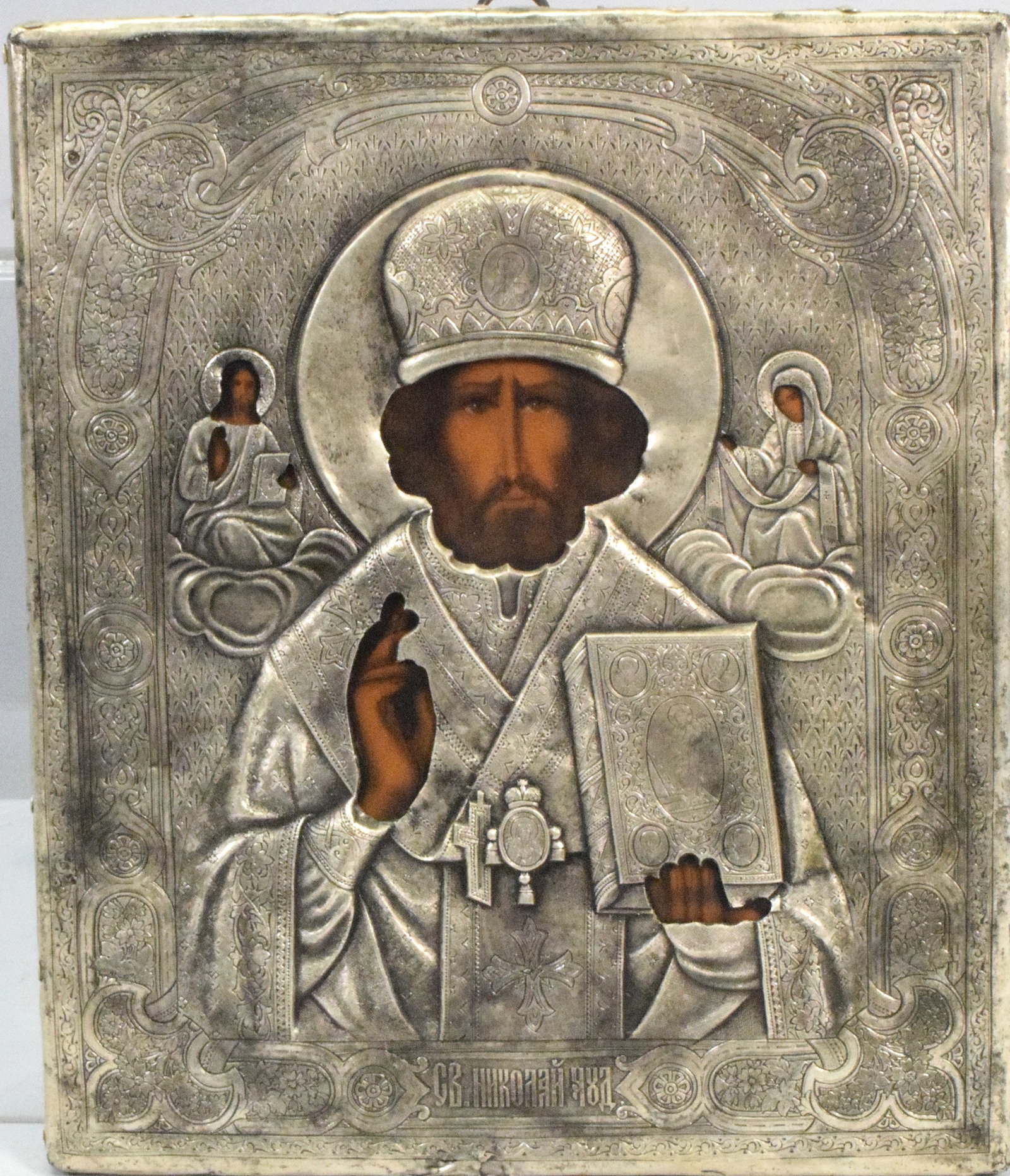 RUSSIAN EMBOSSED SILVER ICON DEPICTING ST NICHOLAS: Finely done Russian Icon of Saint Nichols, rendered in repusse silver or silver plate with a hand painting wooden face and hands. Featuring intricate detailing and Cyrillic incription. Ca19th century.