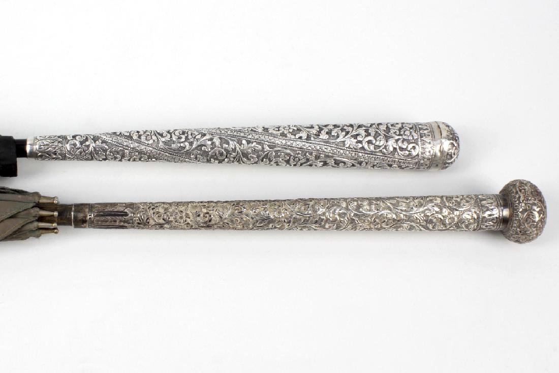 Two Eastern white metal-handled parasols or umbrellas.: Two Eastern white metal-handled parasols or umbrellas. The first having a turned tapering handle with repousse foliate scrollwork decoration, to an ebonised shaft, the canopy (A/F) marked to ribs PARA
