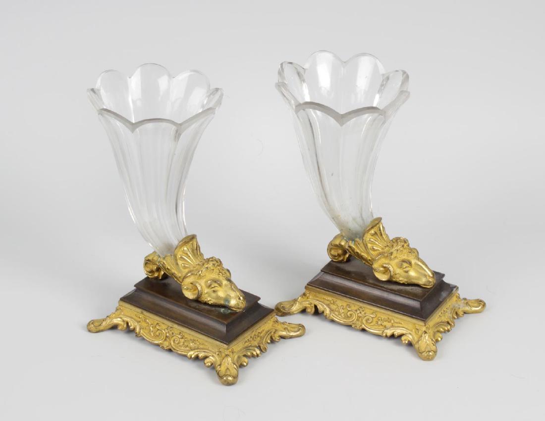 A pair of Regency bronze and ormolu epergnes. Each of: A pair of Regency bronze and ormolu epergnes. Each of cornucopia design with facetted curved glass trumpet vase to a gilt ram's head terminal, on bronze platform and foliate scroll-edged gilt apron wi