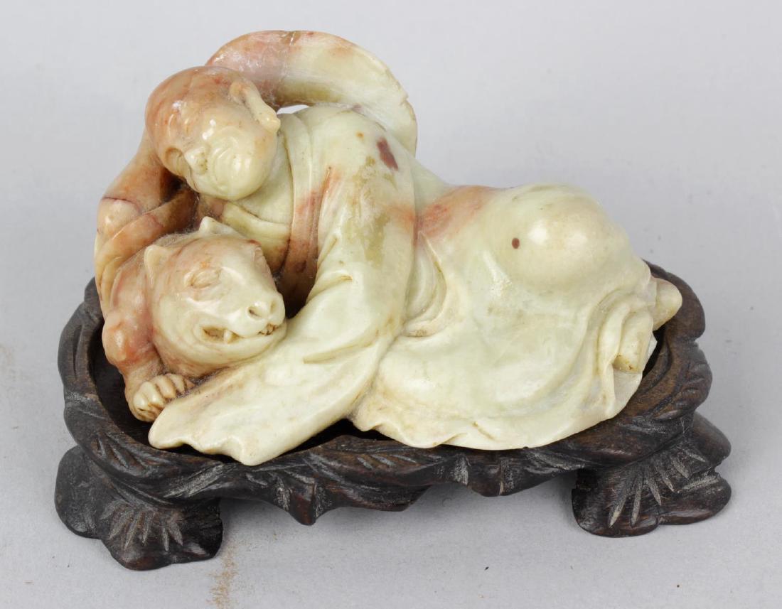 A Chinese carved soapstone figure of a sleeping saint.: A Chinese carved soapstone figure of a sleeping saint. Modelled in recumbent pose with halo (s/d), a dog tucked under his arm, 3, (7.5cm) wide, on original fitted hardwood stand, 2, (5cm) high overall