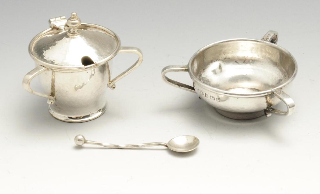 An Arts & Crafts style silver open salt and mustard pot: An Arts & Crafts style silver open salt and mustard pot with spoon, each of circular planished form with three curved handles, the mustard pot opening to reveal a blue Ruskin pottery liner, the spoon 