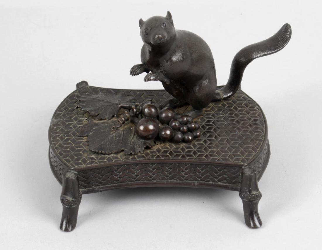 A Japanese Meiji period bronze, modelled as a rat: A Japanese Meiji period bronze, modelled as a rat upstanding on rear legs, poised above a small bunch of grapes, the oblong faux wicker work base upon bamboo form legs, 5.25 (13.25cm) wide.