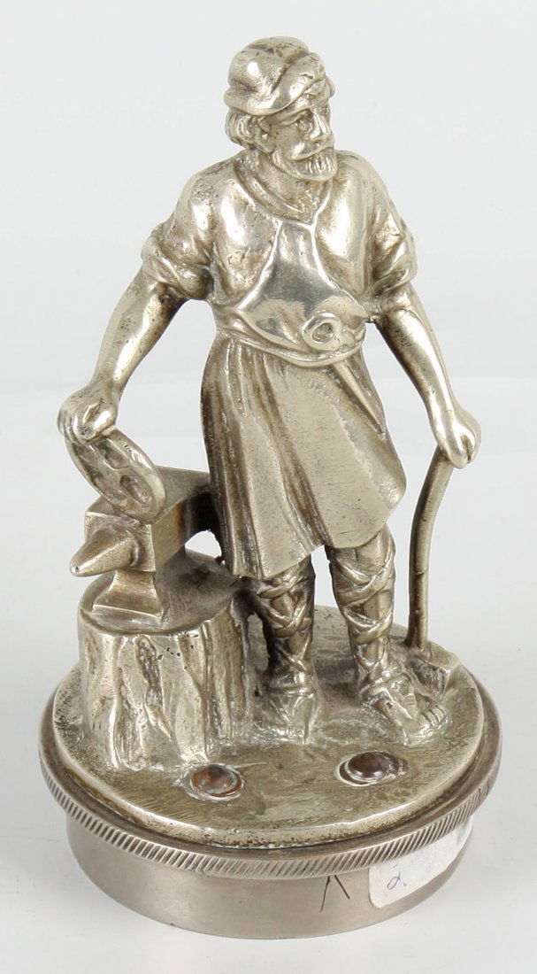 A chromed cast metal automobile radiator mascot: A chromed cast metal automobile radiator mascot modelled as a blacksmith standing near an anvil, holding a wheel and a hammer in his hands. The whole upon a naturalistic modelled base, attached to the