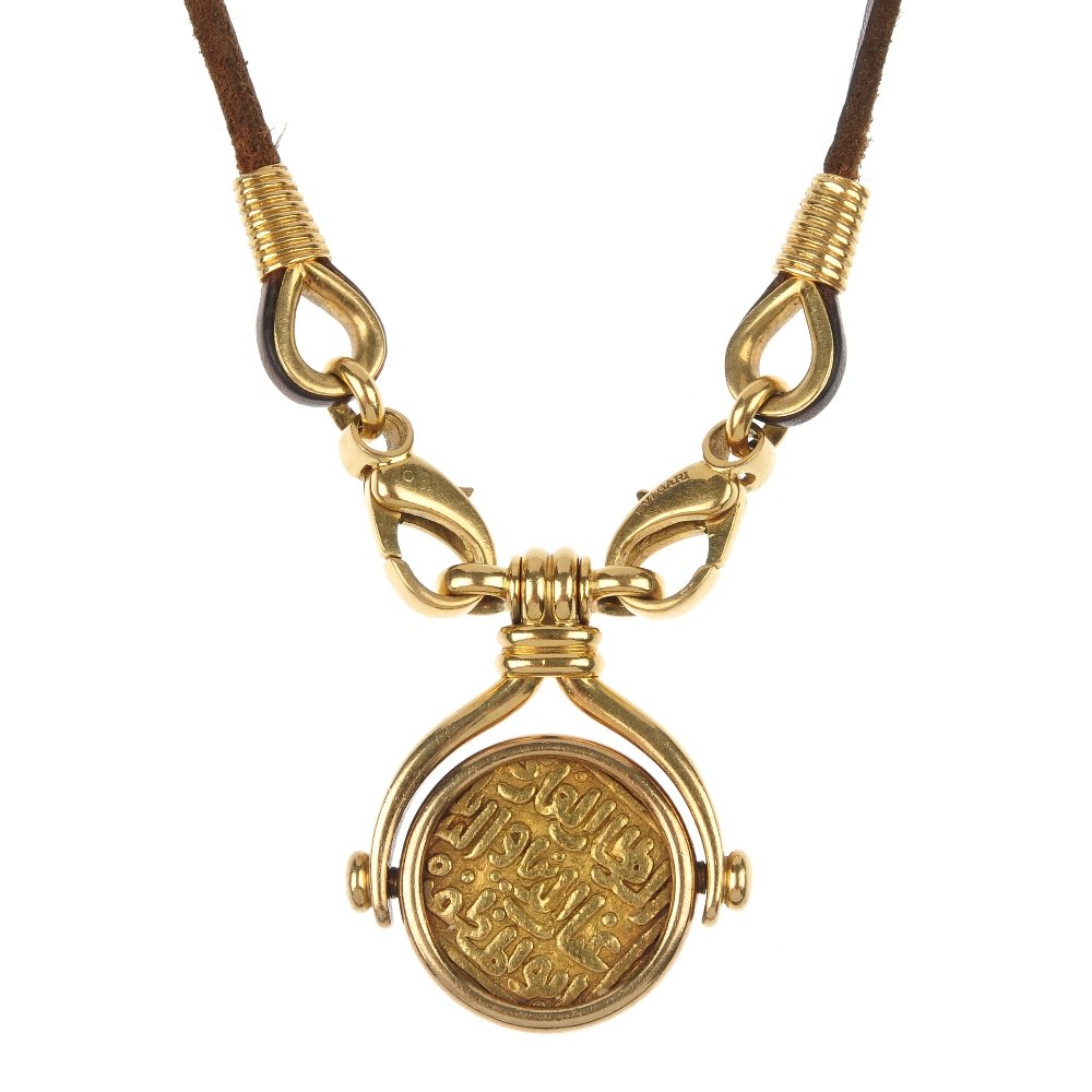 BULGARI - a coin pendant.: BULGARI - a coin pendant. Designed as a rotating mounted coin with embossed Arabic inscription, to the curved bar sides and ridged surmount, suspended from two lobster clasps and a brown leather cord