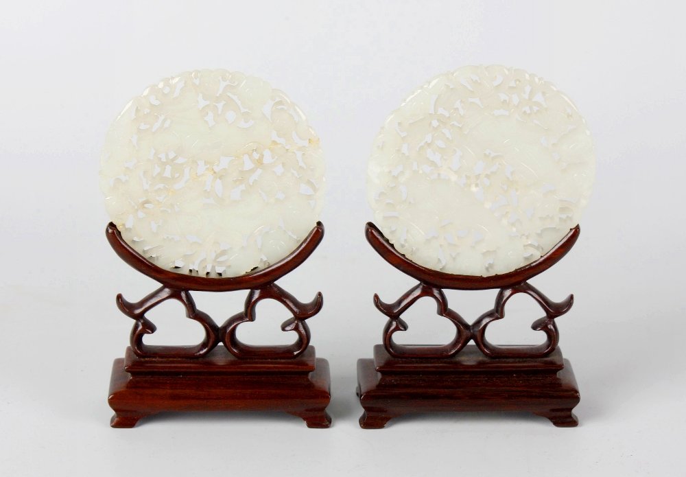 A pair of carved and pierced circular mutton fat jade: A pair of carved and pierced circular mutton fat jade panels, each decorated with figures within scrolled and floral borders, mounted upon a carved hardwood stand, 3" (7.5cm) diameter.