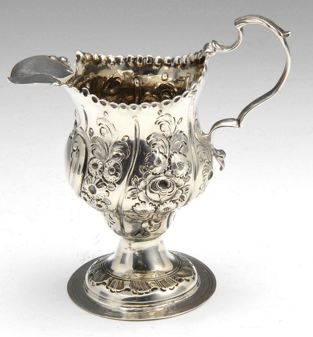 A George III silver cream jug.: A George III silver pedestal cream jug, with simple punched rim above floral and foliate embossed decoration surrounding a vacant cartouche, with foliate capped scroll handle. Hallmarks partly distort