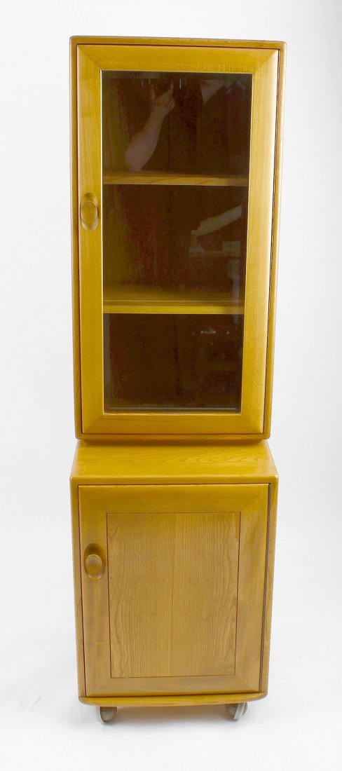 An Ercol light oak two-stage cabinet: An Ercol light oak two-stage cabinet The glazed upper section over projecting cupboard base, 18" x 17" x 64" high, (46cm x 43cm x 162cm)