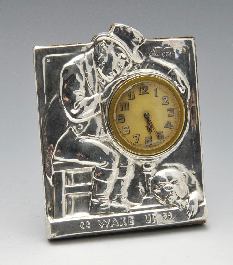 An Edwardian silver mounted novelty bedside clock of: An Edwardian silver mounted novelty bedside clock of rectangular form with wooden easel back, the silver front modelled with a sleeping man seated on a chair and leaning over the clock face with a bul