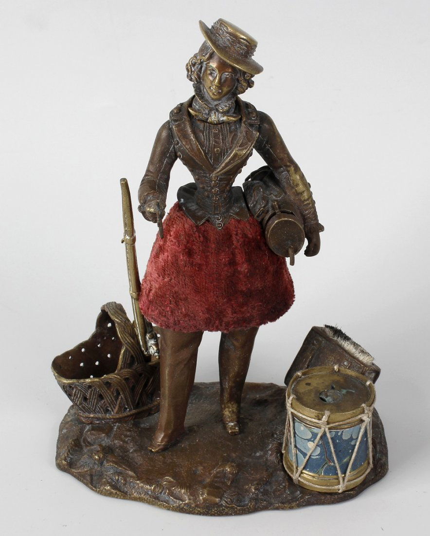 An unusual 19th century French bronze desk necessaire: An unusual 19th century French bronze desk necessaire modelled as a lady carrying a rifle and other wares, upon a naturalistic modelled base detailed with basket, drum, etc., her removable head formin