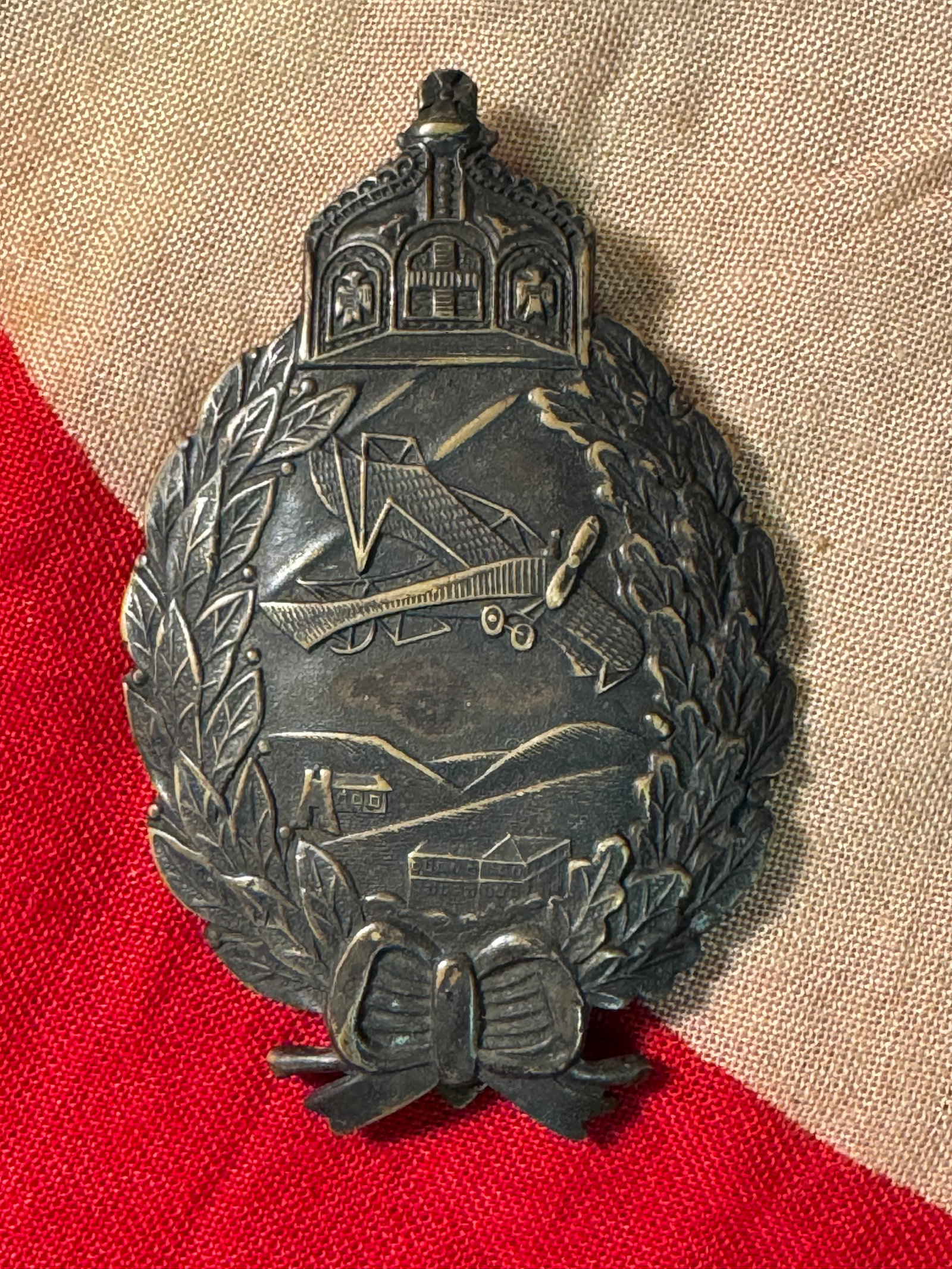 WWI IMPERIAL GERMAN PRUSSIAN PILOT BADGE: WWI IMPERIAL GERMAN PRUSSIAN PILOT BADGE. The front shows a flying airplane in the center with farm houses and hills below. There is a King?s crown at the top with a bow at the bottom. Has an oval oak