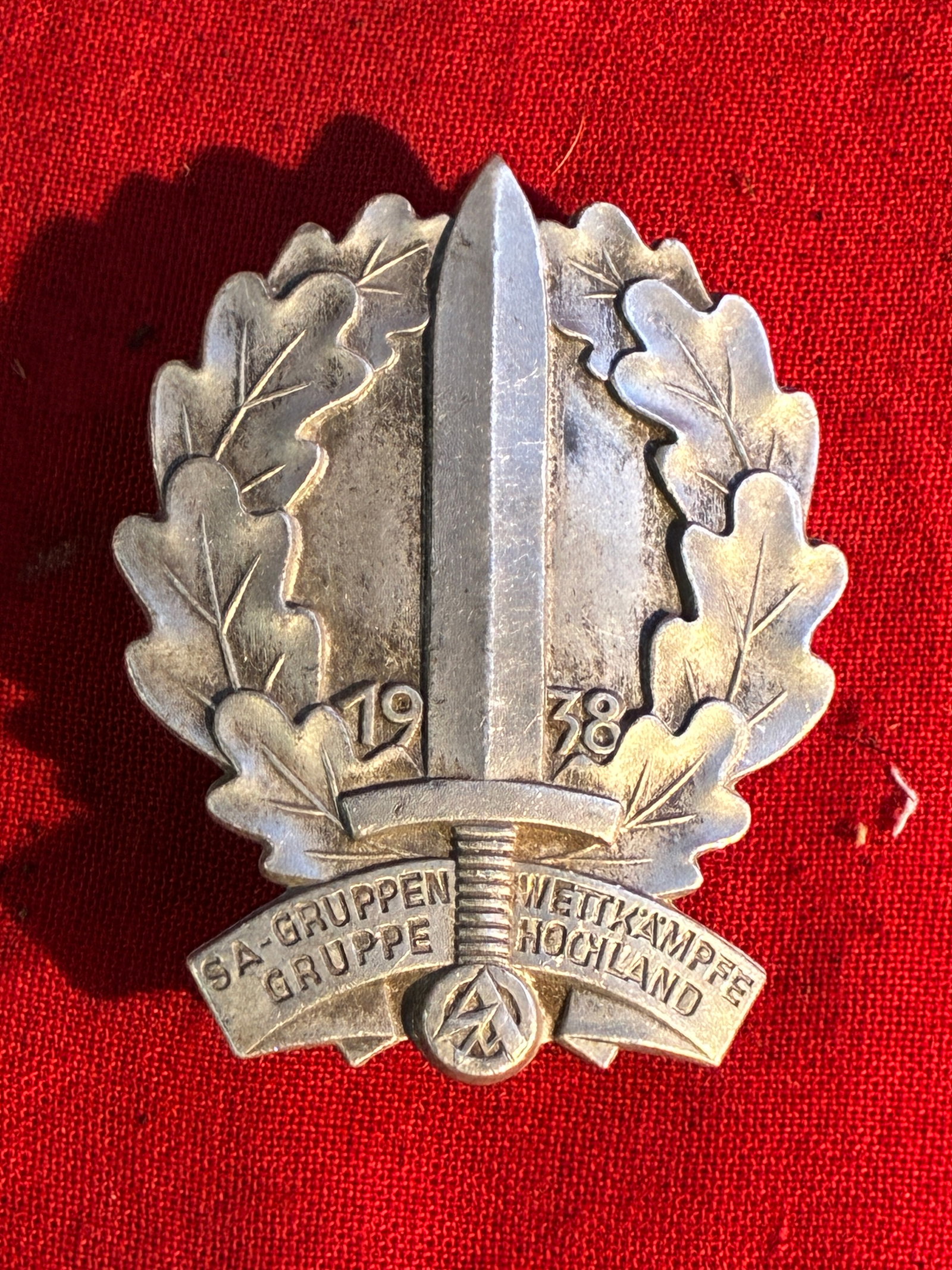 WW2 German NSDAP (NAZI PARTY) BrownShirts 1938 SA GRUPPE HOCHLAND SPORTS COMPETITION BADGE: WW2 German NSDAP (NAZI PARTY) BrownShirts 1938 SA GRUPPE HOCHLAND SPORTS COMPETITION BADGE. An approximately 50mm x 40mm, silver proto-plastic badge. The badge depicts a sword with an SA insignia to i