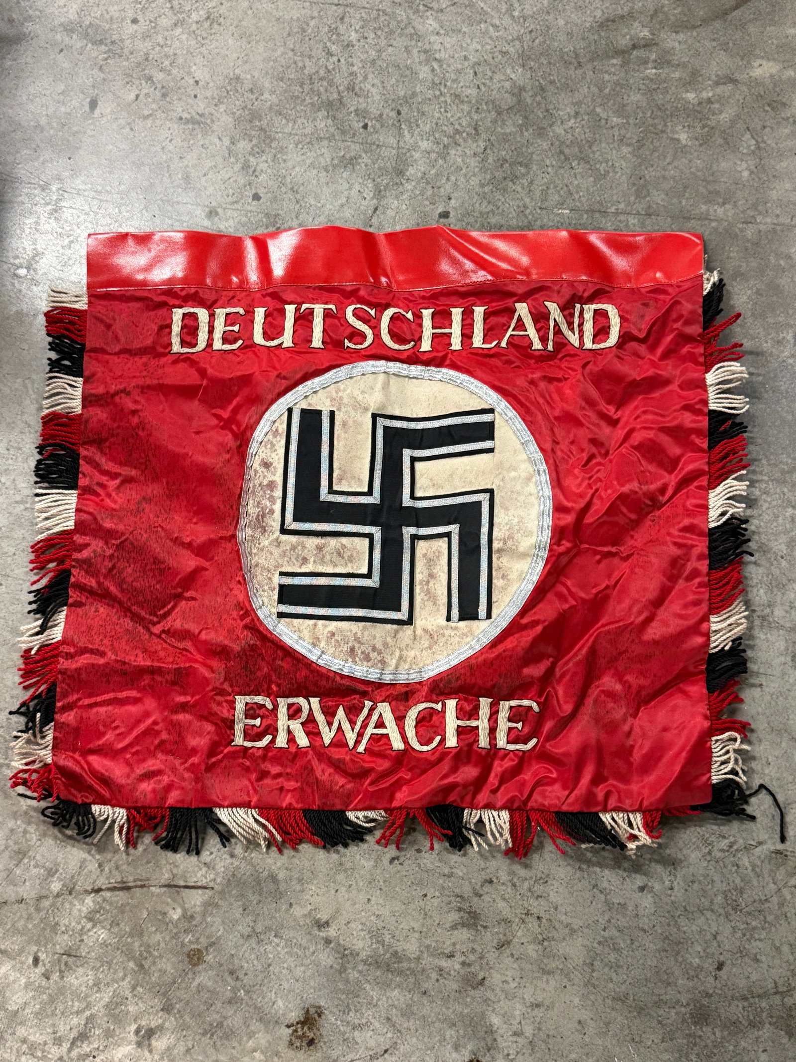 WW2 German NSKK DE Standard Flag: WW2 German NSKK Deutschland Erwache Standard. The front top of the flag says DEUTSCHLAND ERWACHE. On the back top NAT. SOZ. DEUTSCHE ARBEITERPARTEI with STURMABTEILUNG below the swaz. Very worn condit