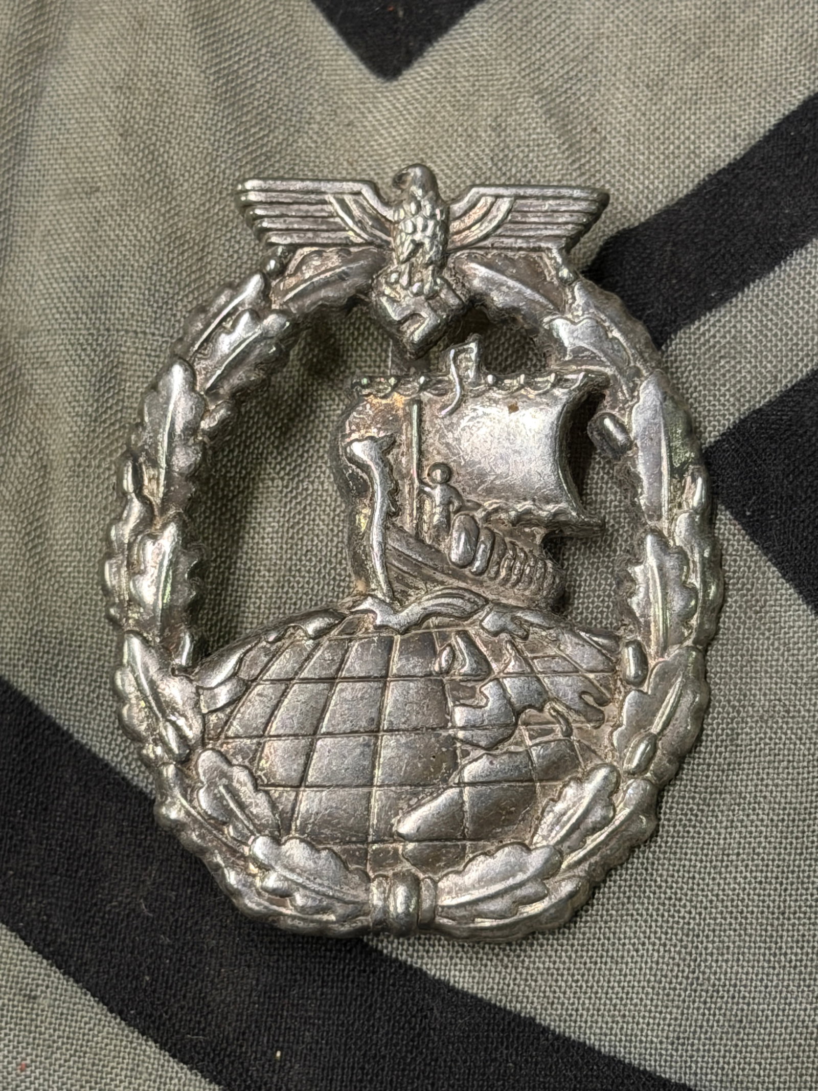 WW2 German Kriegsmarine auxiliary cruiser badge By JMME Berlin: WW2 German Kriegsmarine auxiliary cruiser badge By JMME Berlin. A very rare example. Late war zinc construction with riveted tombac globe. Typical later period S&L production with magnetic setup. Good