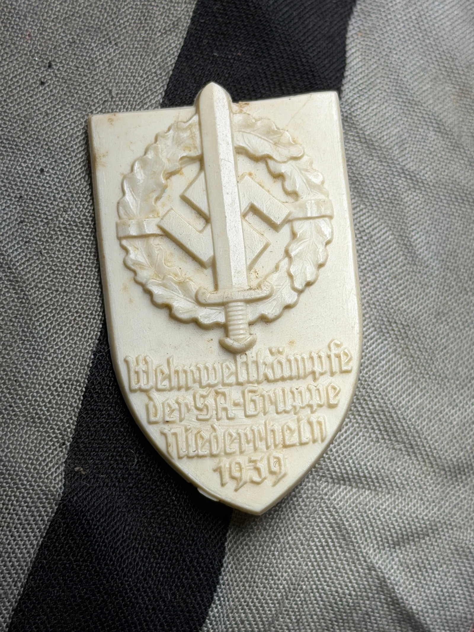 WW2 German NSDAP (NAZI PARTY) Brownshirts 1939 SA Group Niederrhein Sport Day Badge: WW2 German NSDAP (NAZI PARTY) Brownshirts 1939 SA Group Niederrhein Sport Day Badge. 35mm x 50mm, white protoplastic badge with bronze coloured paint. The badge depicts a sword, with an SA insignia to