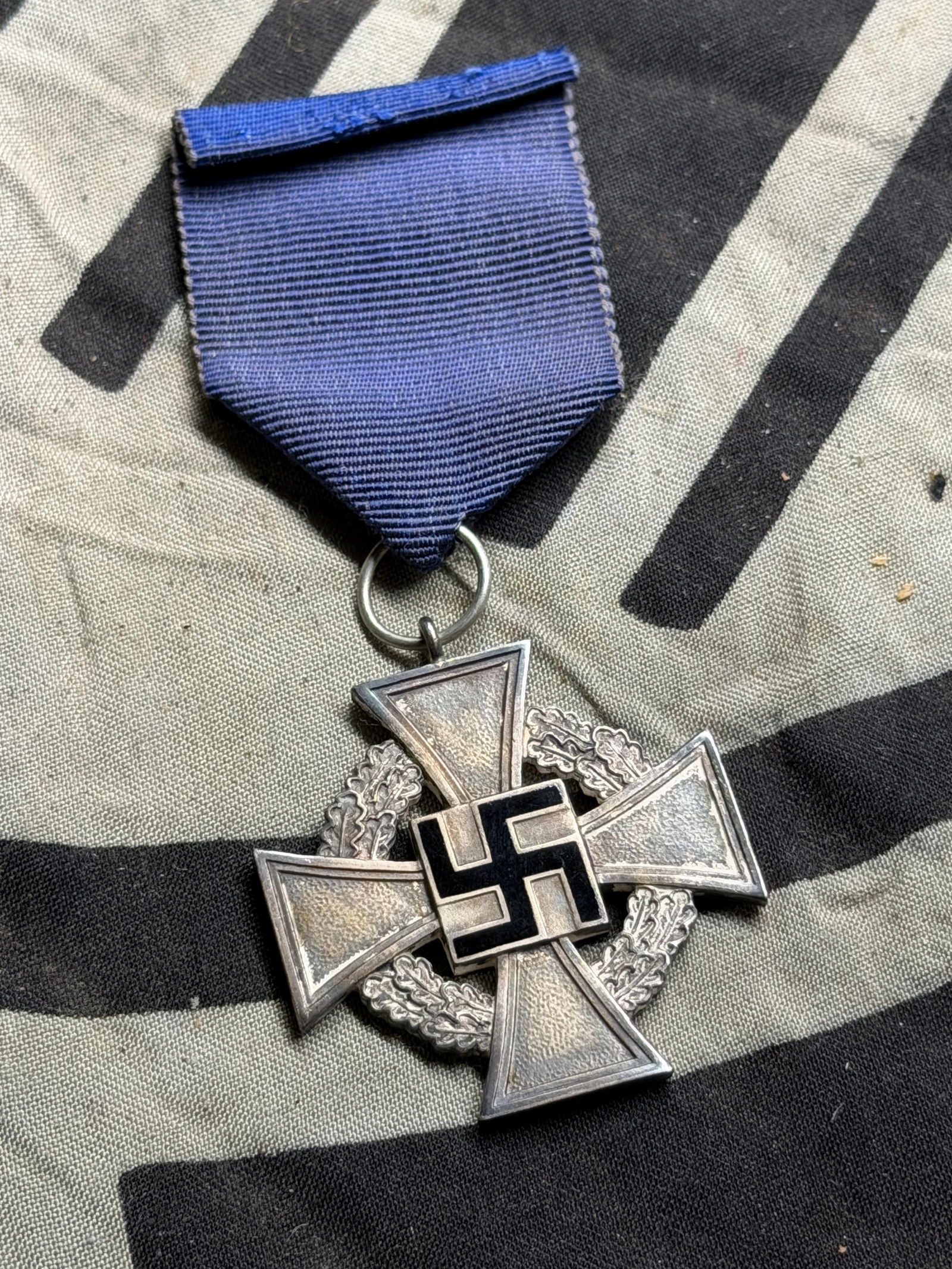 WW2 GERMAN CROSS OF HONOUR FAITHFUL SERVICE CROSS WITH RIBBON: WW2 GERMAN CROSS OF HONOUR FAITHFUL SERVICE CROSS WITH RIBBON. Front: The medal has a Maltese-style cross with a black enameled swastika in the center, surrounded by a silvered wreath of oak leaves. R