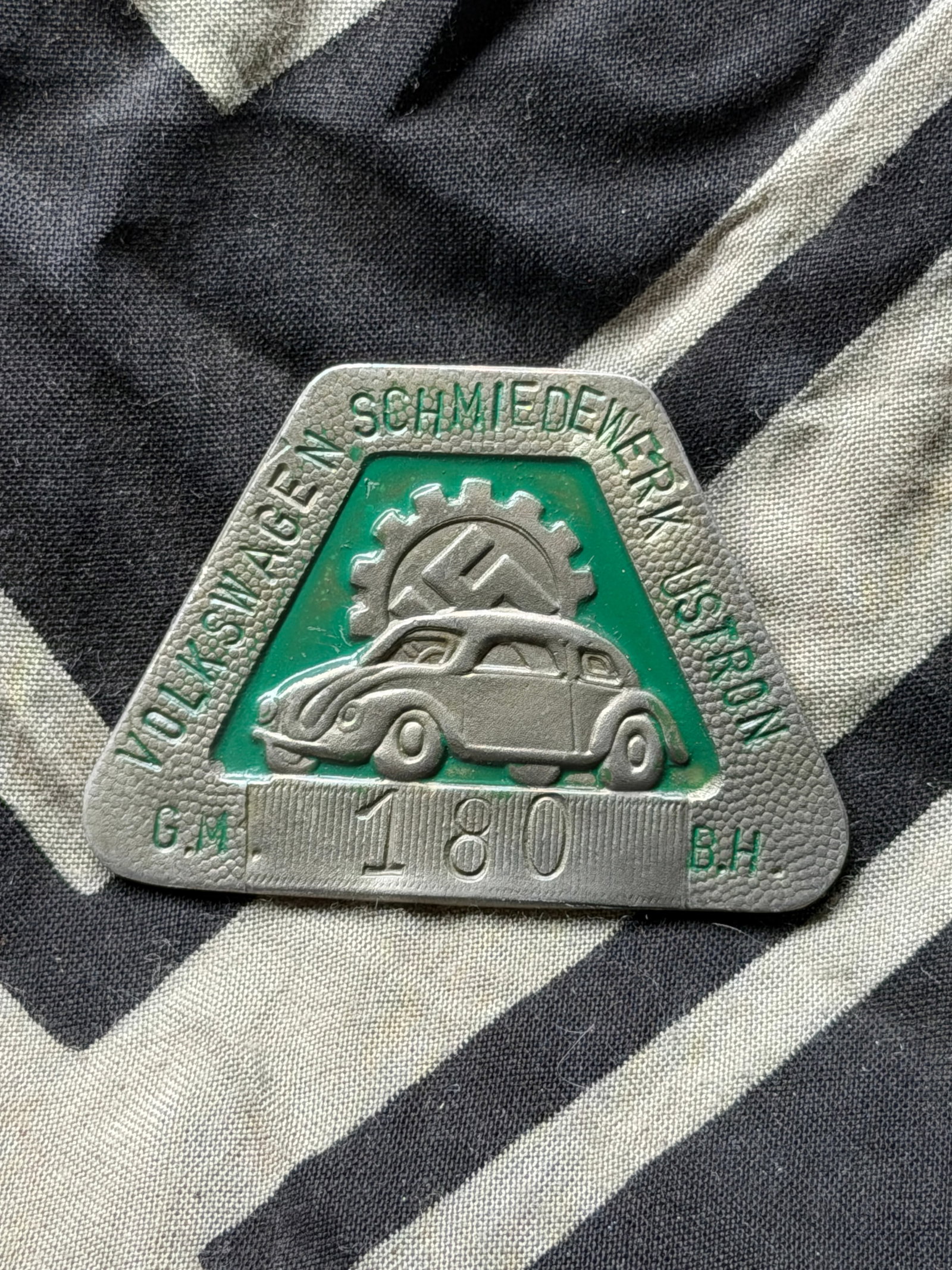 WW2 German Volkswagen Schmiedewerk Ustron Employee Badge: WW2 German Volkswagen Schmiedewerk Ustron Employee Badge. This triangular metal badge features a relief depiction of an early Volkswagen “Beetle” automobile over a green enamel background, sur