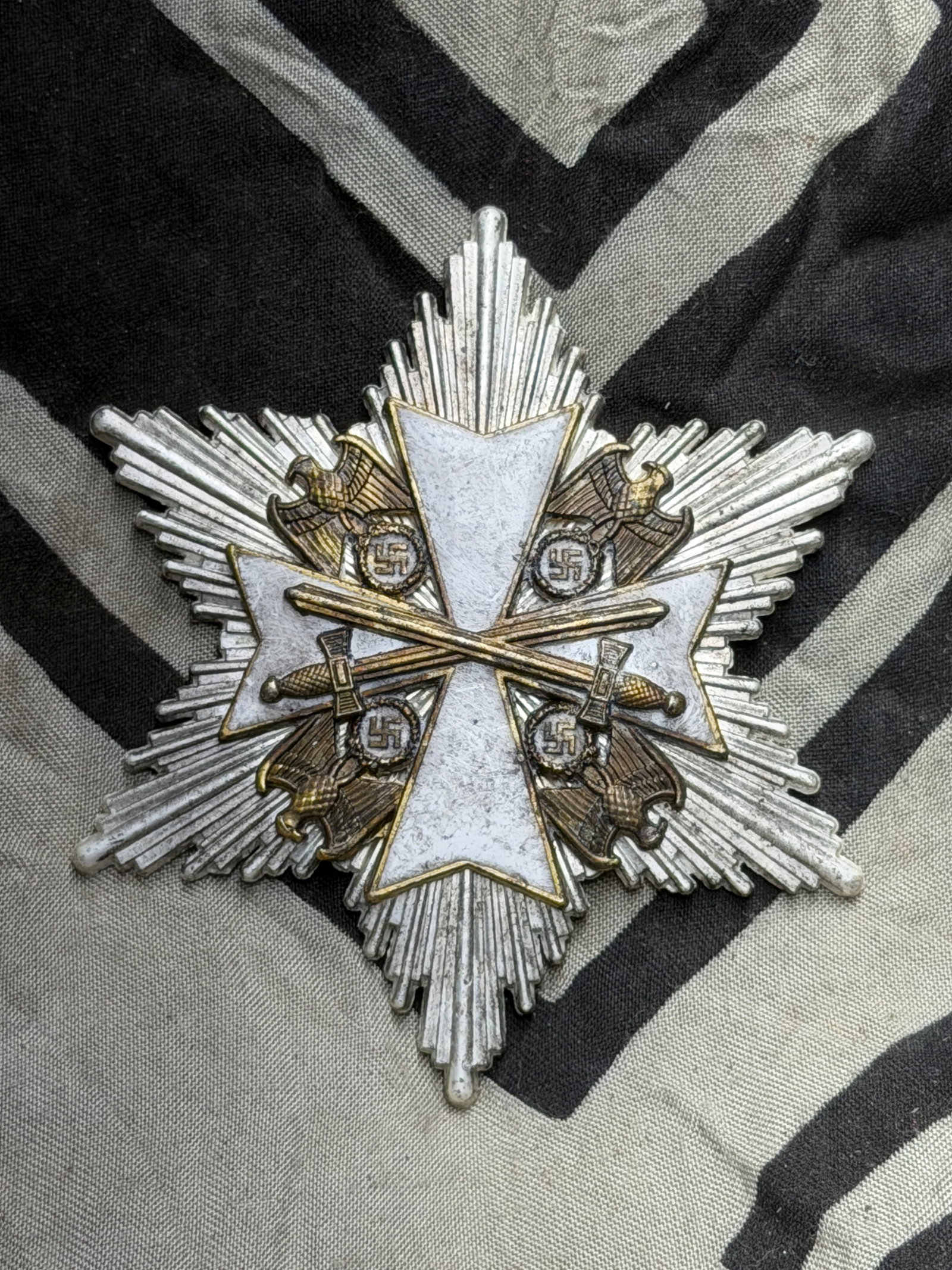 WW2 German War Merit Cross with Swords, Star Form, circa 1940s: This metal insignia is a star-shaped variant of the Kriegsverdienstkreuz (War Merit Cross), an award instituted during the Second World War. It features a white-enamel cross over a silver radiating st