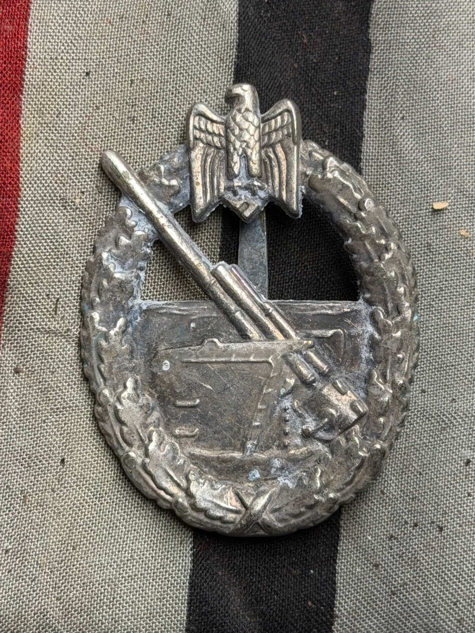 General Assault Badge (Allgemeines Sturmabzeichen), WW2 Germany, circa 1940s: This silver-toned metal badge depicts an oval wreath of oak leaves surrounding a crossed bayonet and stick grenade over a German eagle clutching a swastika. The reverse bears a vertical pin with a hin