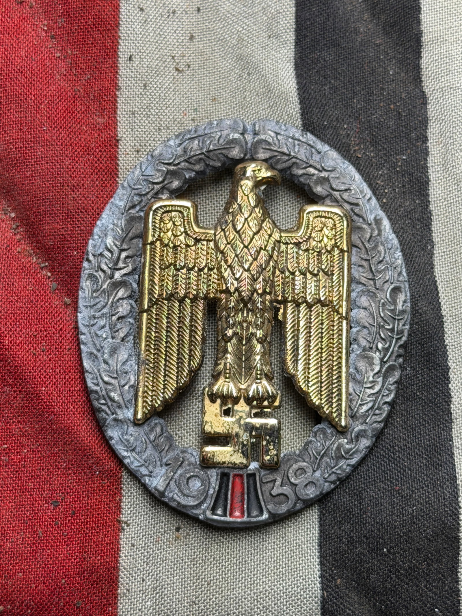 Commemorative Badge — “Anschluss Medal?? (Medaille zur Erinnerung an den 13. März 1938), WW2: This oval metal badge features a gilt eagle grasping a swastika, set within a silvered wreath of oak leaves. At the base is the date “1938” above a small red, white, and black enamel panel rep