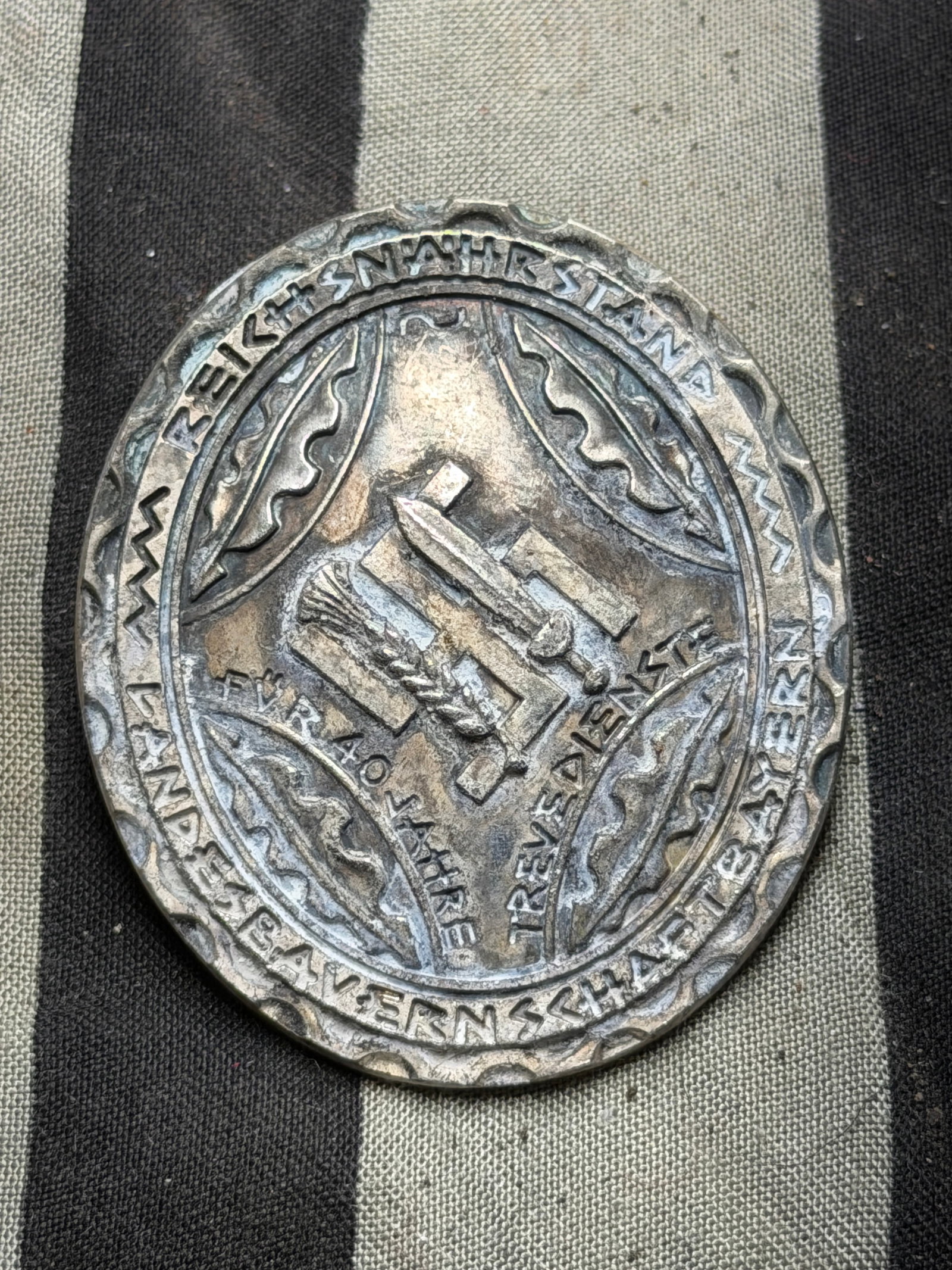 Reichsnährstand 40-Year Service Badge — Bavarian Farmers’ Association, WW2 Germany,: This circular, silver-toned metal badge features a central motif of a plow, sword, and wheat sheaf arranged in a triangular composition, symbolizing agriculture, defense, and labor. Around the perimet