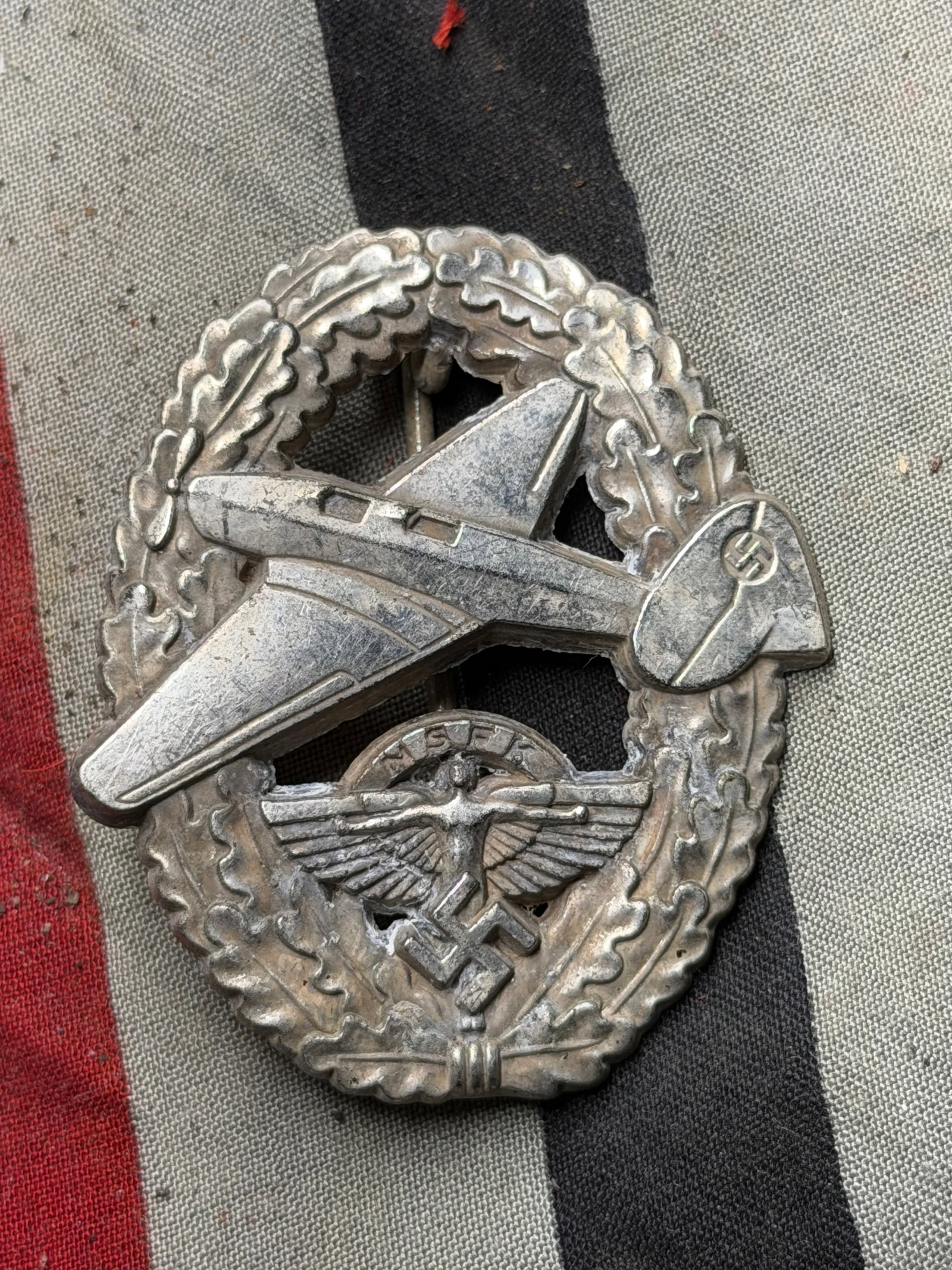 WW2 German NSFK Glider Pilot Badge (National Socialist Flying Corps), c. 1930s–1945: This silver-toned metal badge features a glider aircraft in flight, superimposed over a wreath of oak leaves, with the emblem of the National Socialist Flyers Corps (Nationalsozialistisches Fliegerkor