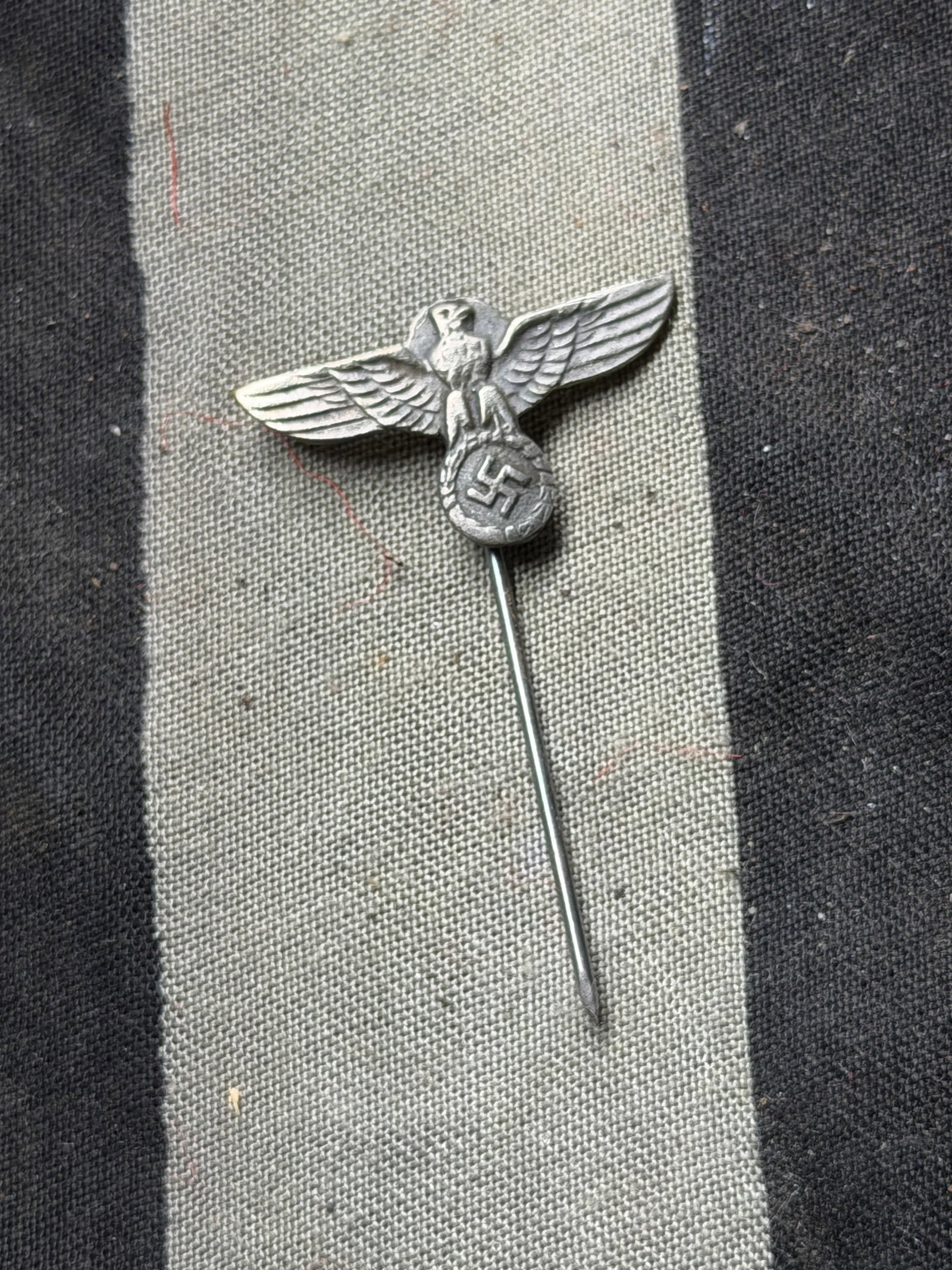 WW2 German National Emblem Stickpin (Eagle and Wreath Motif, Early 20th Century): This small metal stickpin features an eagle with outstretched wings, clutching a wreath enclosing a swastika—a symbol used in Germany during the 1930s–1940s. Such emblems were employed by various 