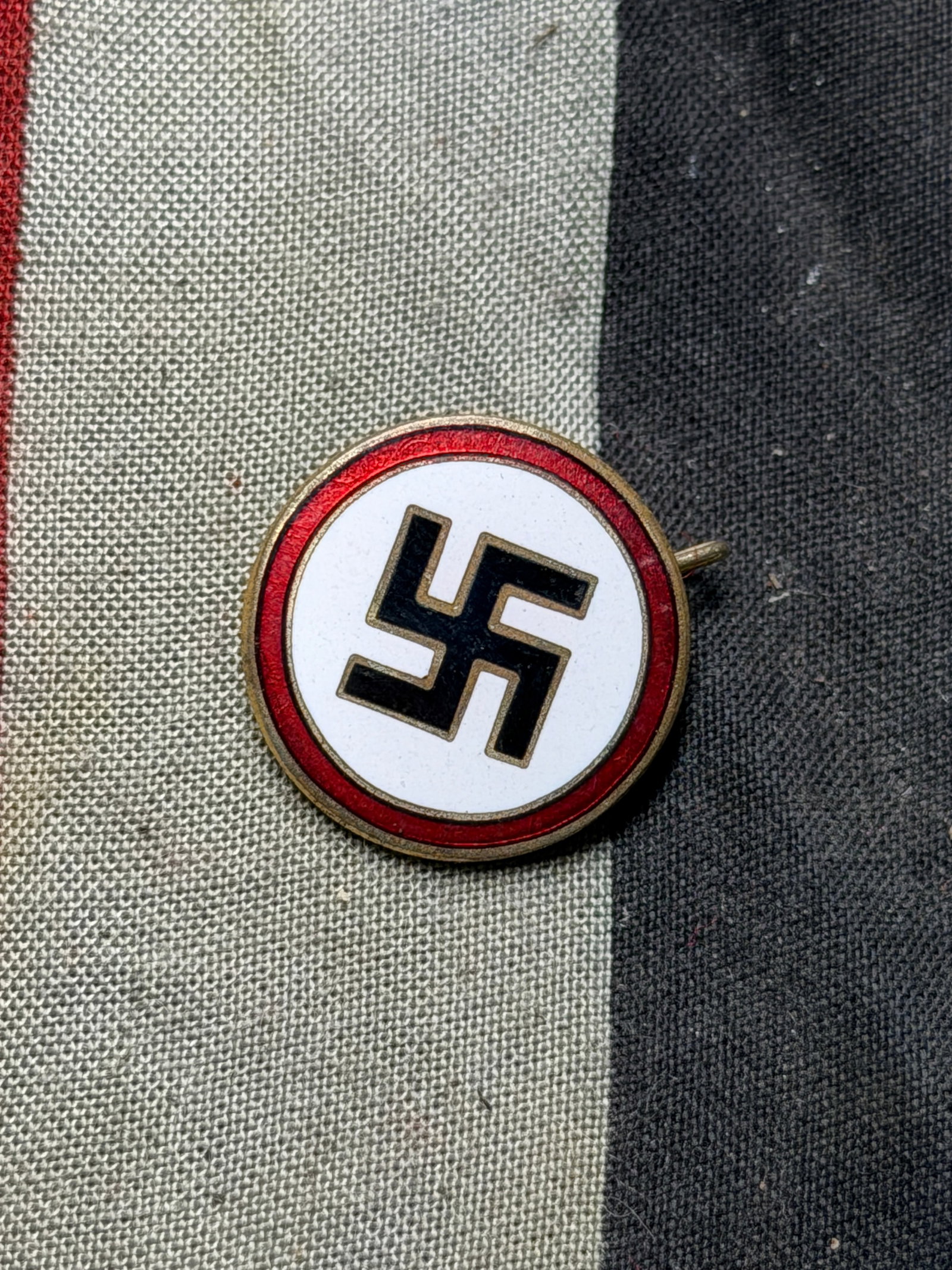 WW2 German Political Party Membership Badge (Early 20th Century): A circular enameled metal badge featuring a red, white, and black design with a central hooked cross symbol, historically used in Germany during the 1930s–1940s. The reverse bears a pin-back fastene