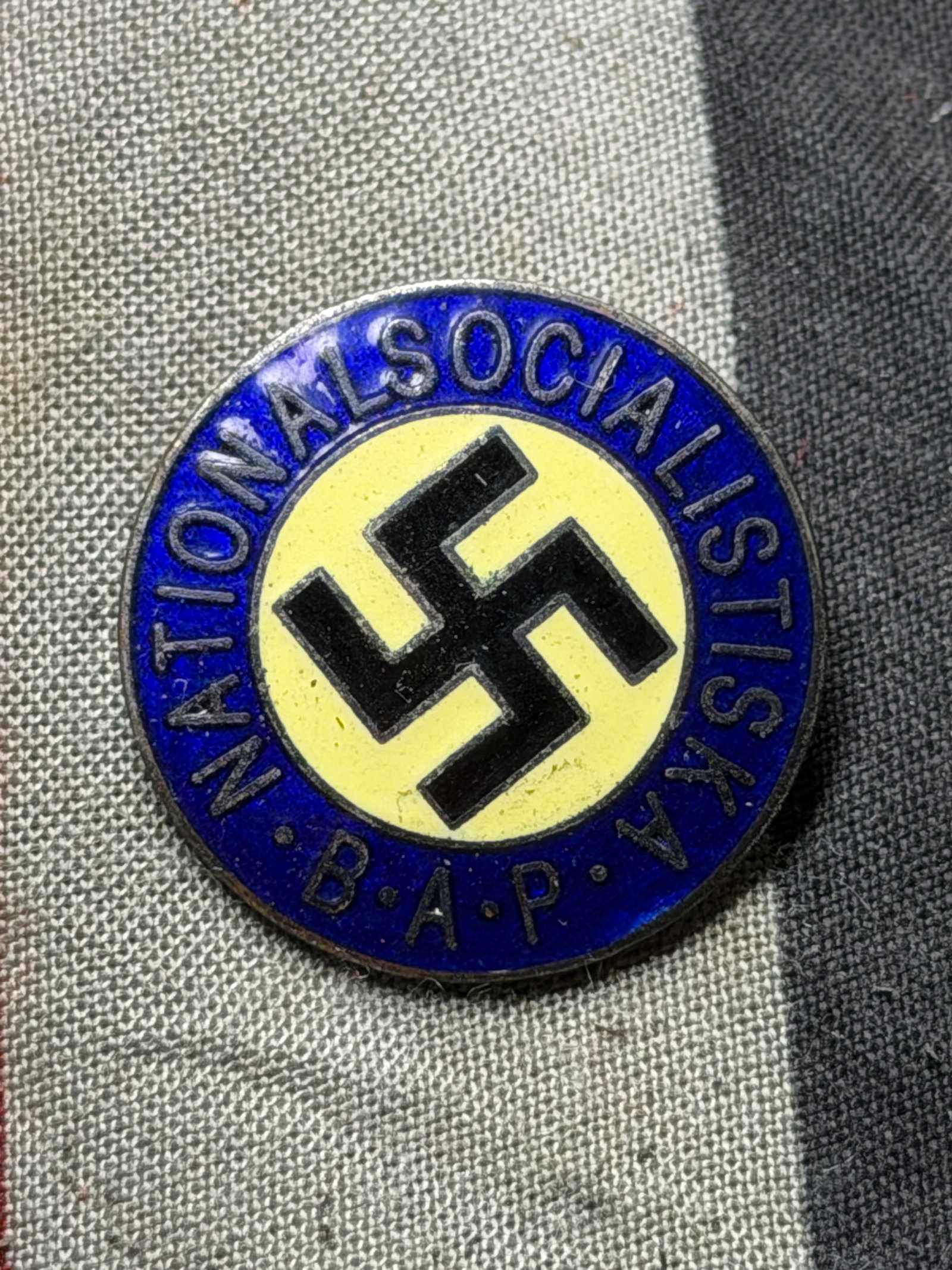 WW2 German Swedish National Socialist Enaeled Badge (Early 20th Century): This is a circular metal and enamel badge featuring the inscription “Nationalsocialistiska B.A.P.” around a central symbol on a yellow and blue background. The reverse side shows a simple pinb
